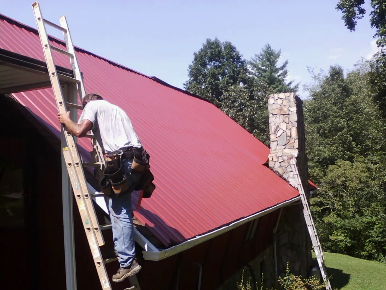 Roofer on a ladder beside a red metal roof and stone chimney — Soffit & Fascia Repair by Roofing Brothers in Danville